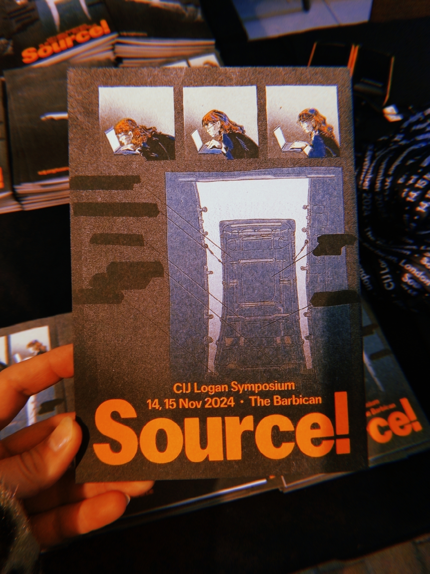 A hand holds a flyer for the CIJ Logan Symposium "Source!" at The Barbican, featuring illustrations and event details.