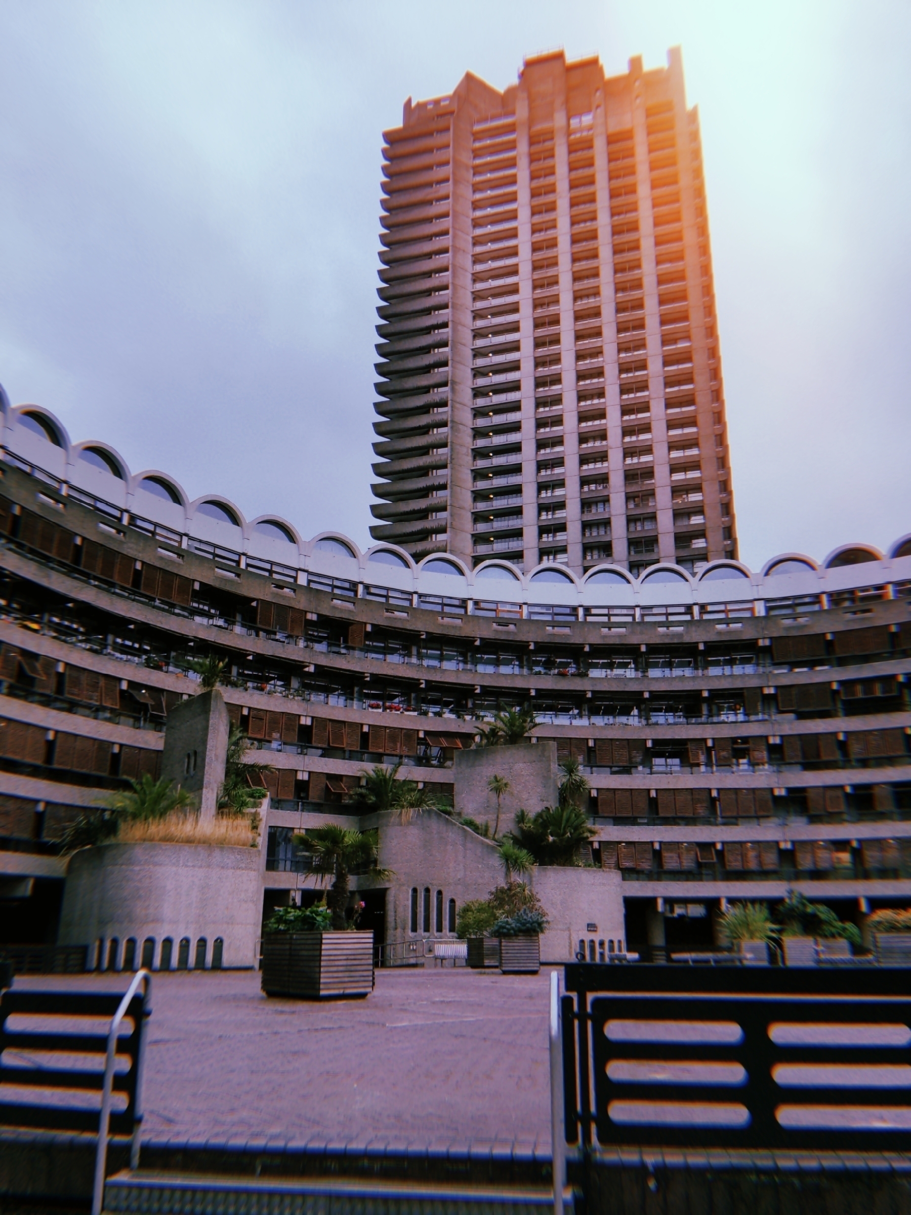 A tall, uniquely structured tower stands behind a curved, modern building with multiple floors and a patterned facade.