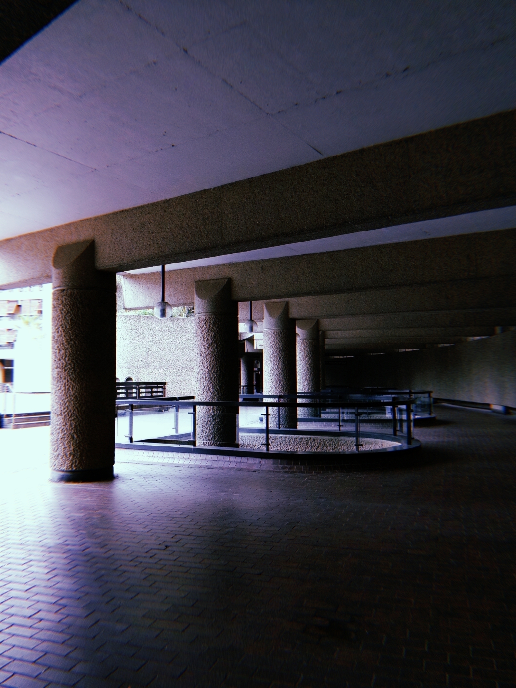 A dimly lit architectural space features textured columns, angular railings, and muted brick flooring.