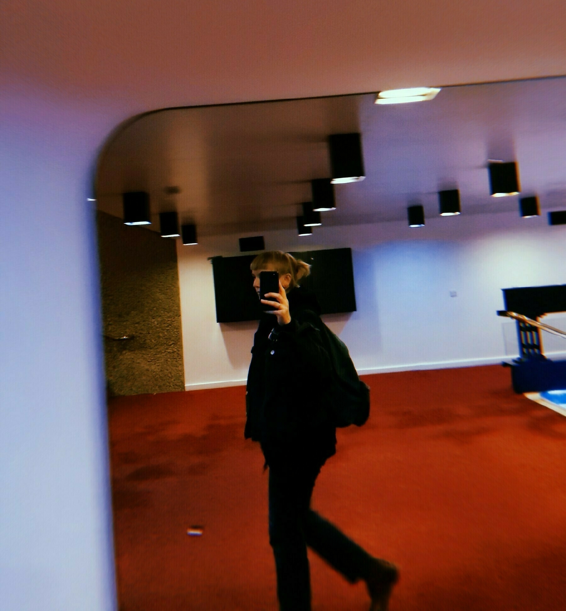 Tansy takes a selfie while walking past a mirror. she is walking in a red-carpeted lobby area with black ceiling lights and a reflective surface on the left.