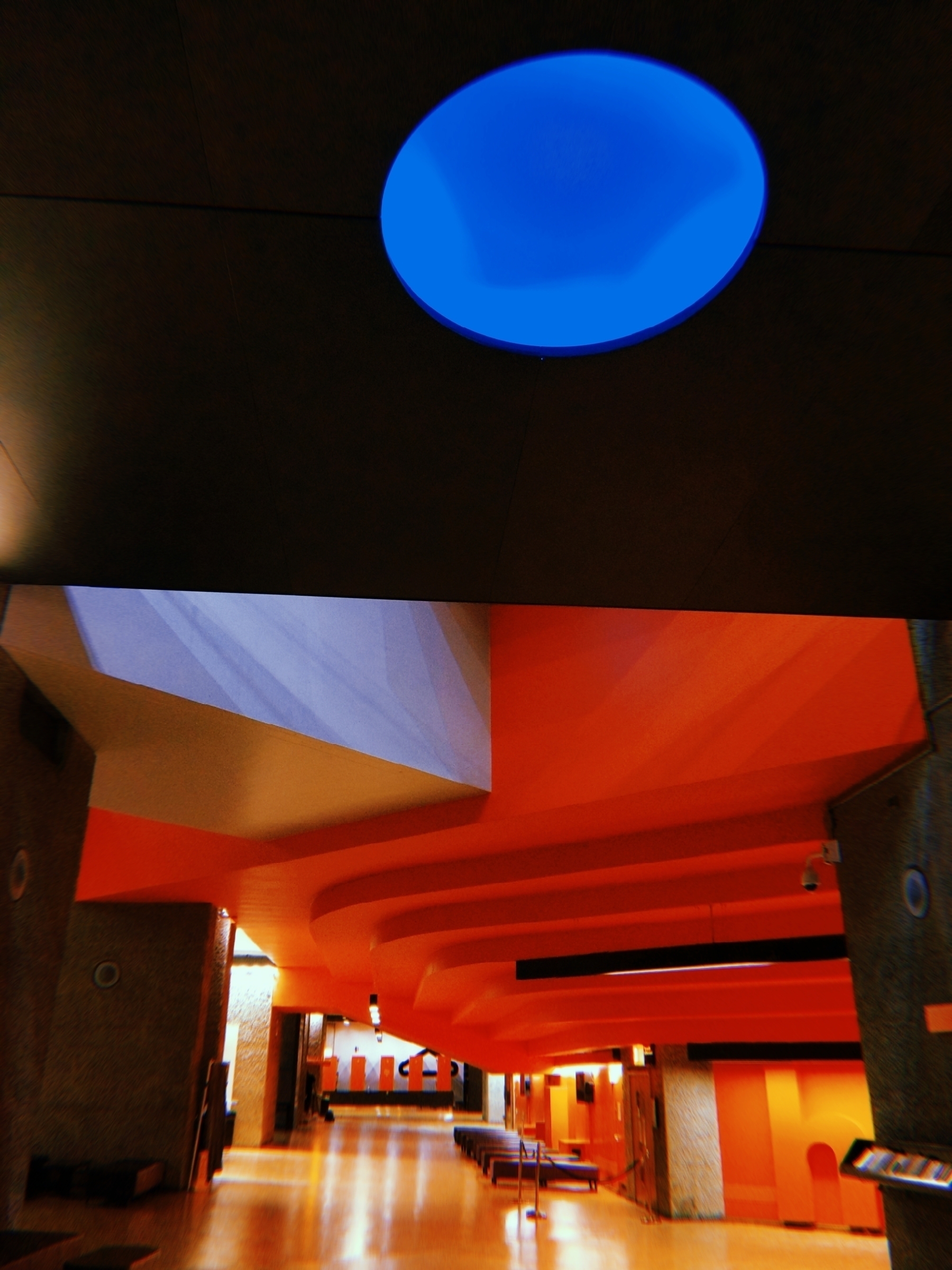 A modern architectural interior features bold orange walls, geometric shapes, and a striking blue circular skylight.