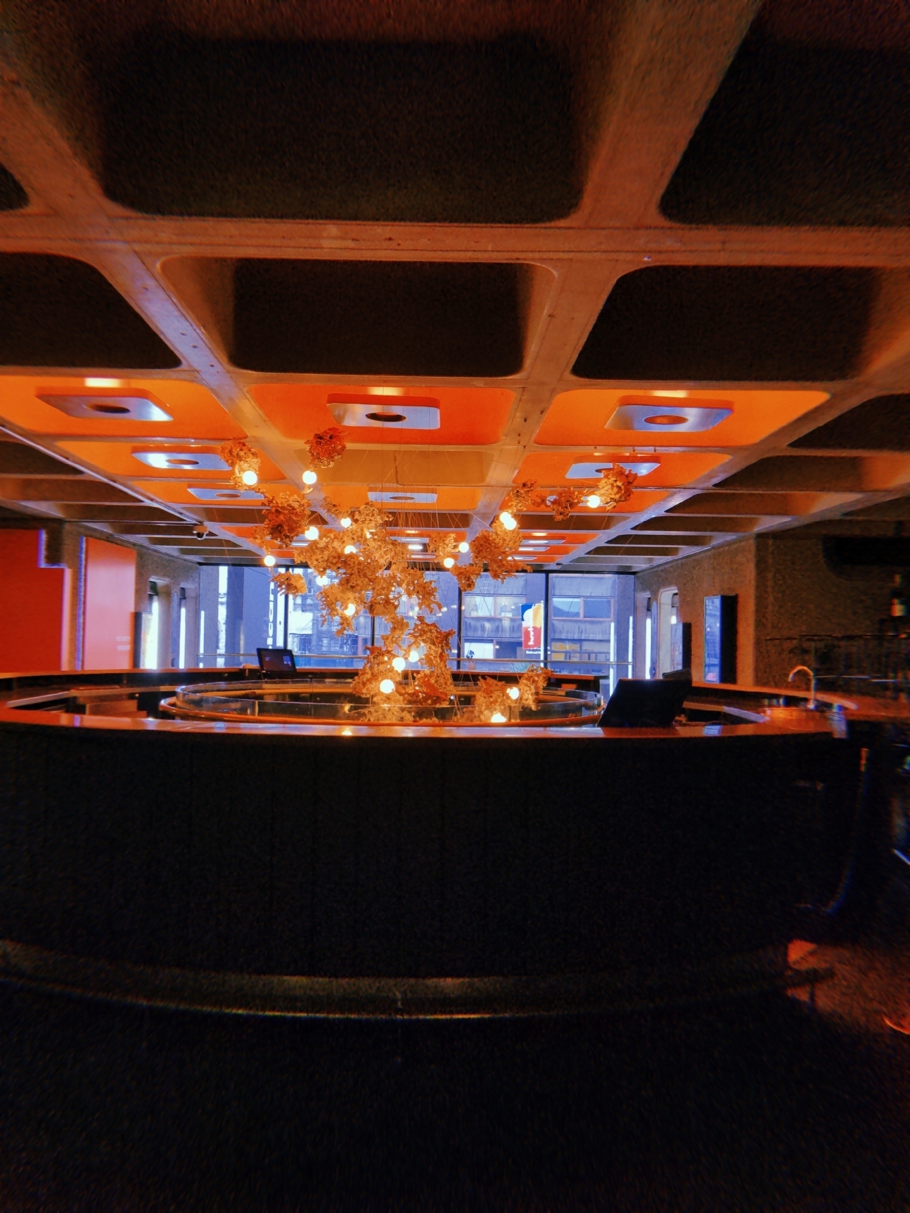 A modern interior space features a circular bar with a unique lighting fixture composed of hanging globes, set against large windows and an illuminated ceiling.