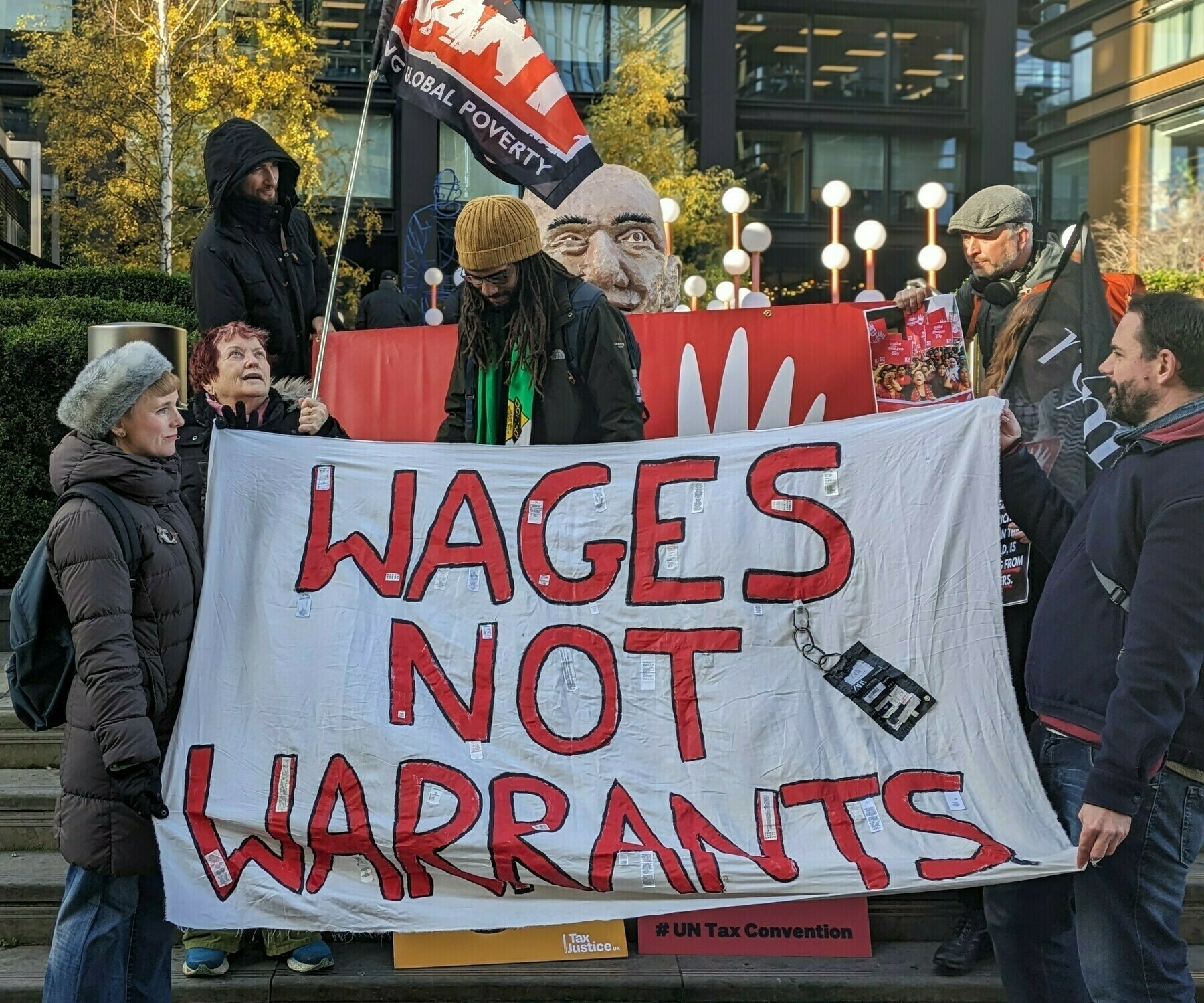 A group of people gathered to protest outside Amazon HQ. Two red and white banners say Make Amazon Pay and Wages Not Warrants. There is a giant sculpture of Jeff Bezos's head