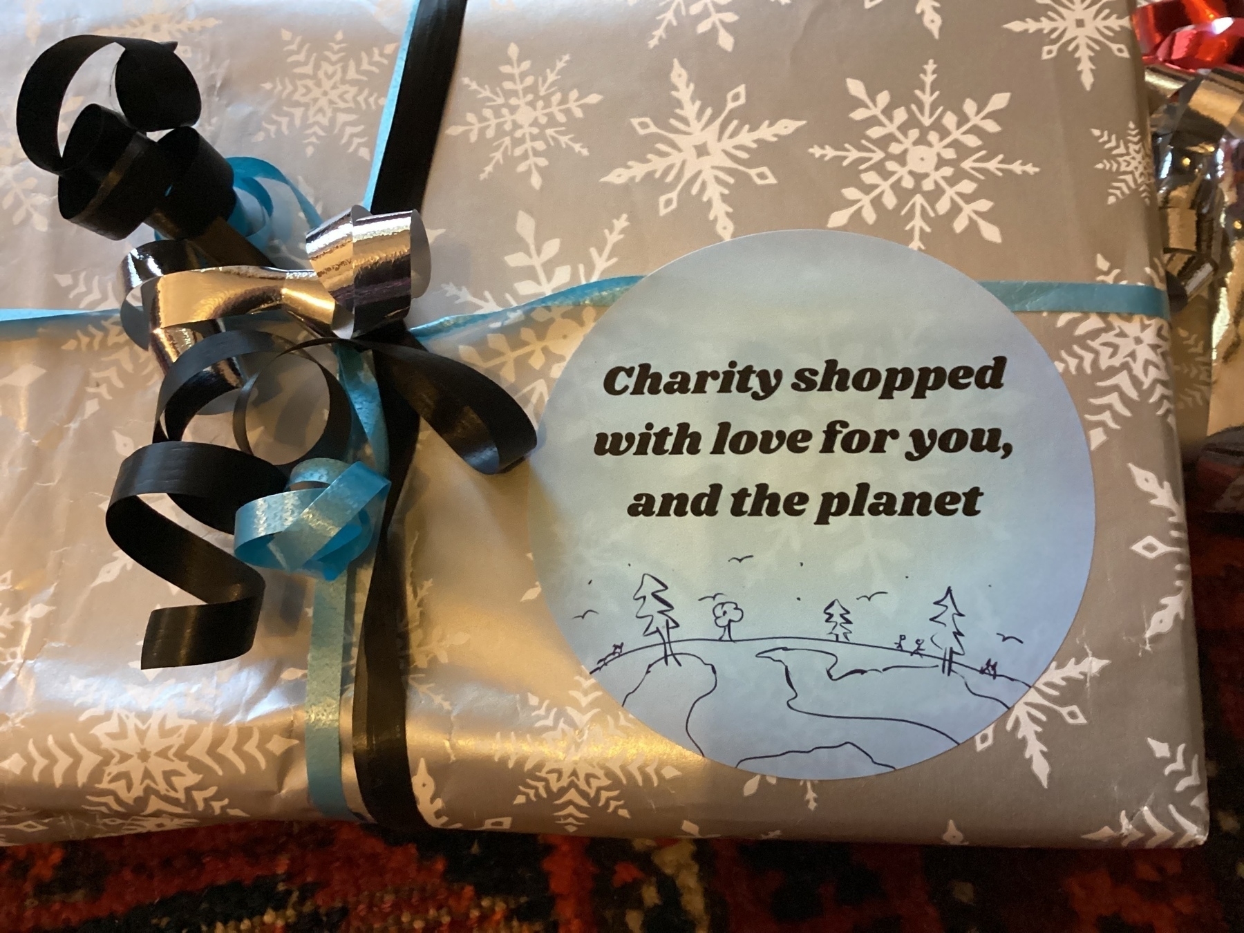 A gift wrapped in silver snowflake paper is tied with blue and black ribbons, featuring a circular label that reads, "Charity shopped with love for you, and the planet."