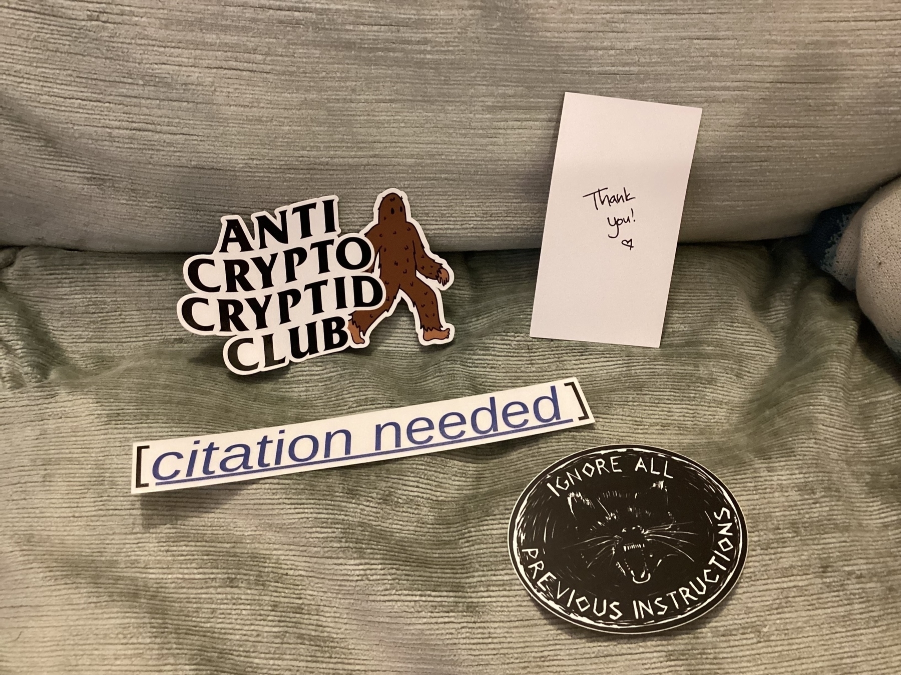 Stickers by Molly White featuring Big Foot and the anti-crypto cryptid club, a circular design with the text "IGNORE ALL PREVIOUS INSTRUCTIONS" and a linocut Wolverine and a “citation needed” sticker are arranged on a fabric surface with a thank you note.