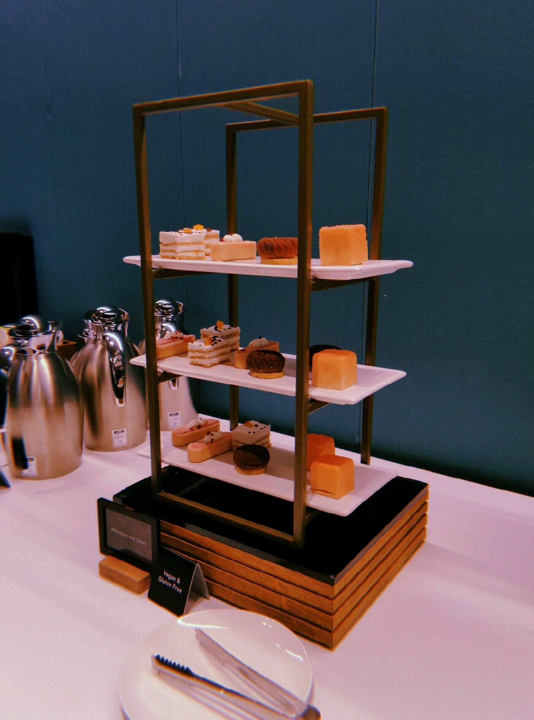 A tiered stand displays an assortment of elegant vegan pastries and treats on a table set with tongs and beverage containers.