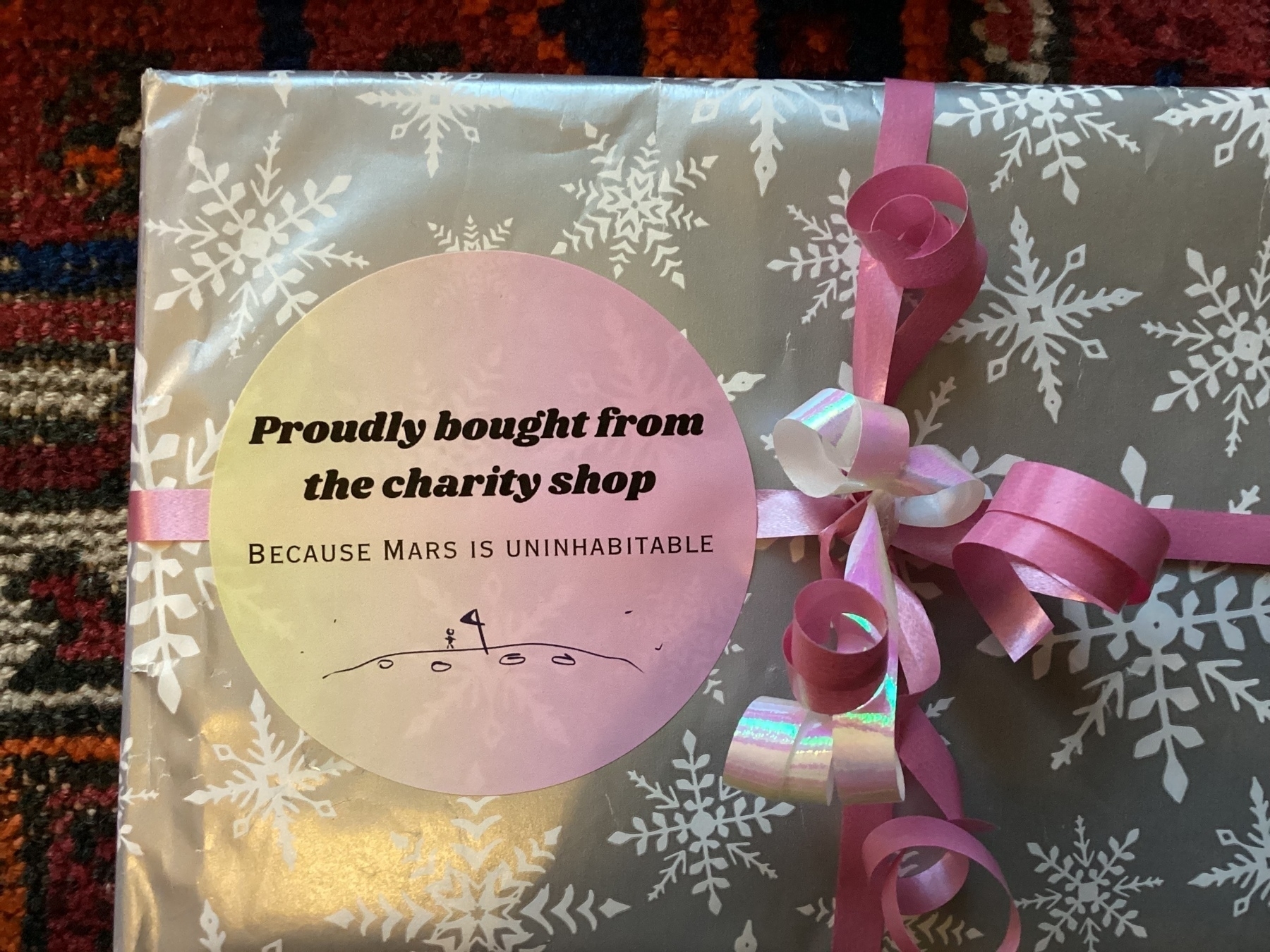 A gift wrapped in snowflake-patterned paper features a humorous sticker that reads, "Proudly bought from the charity shop because Mars is uninhabitable," adorned with pink ribbons.