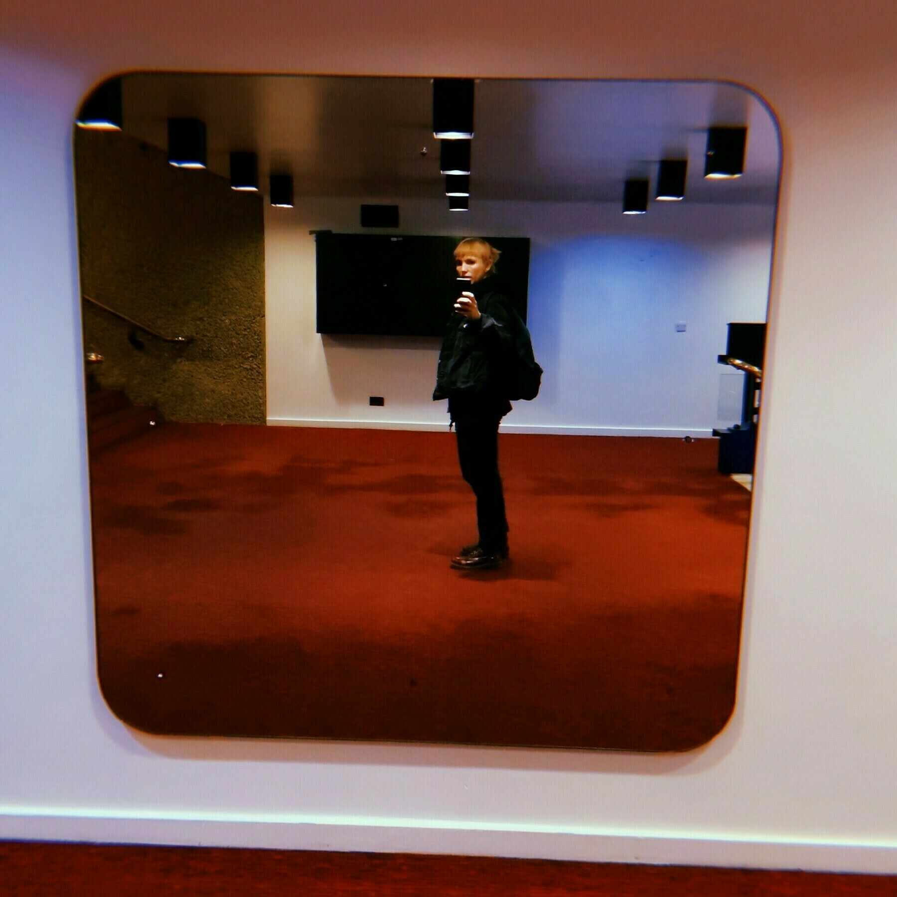 Tansy wearing black is taking a mirror selfie in a room with red carpet and ceiling lights.