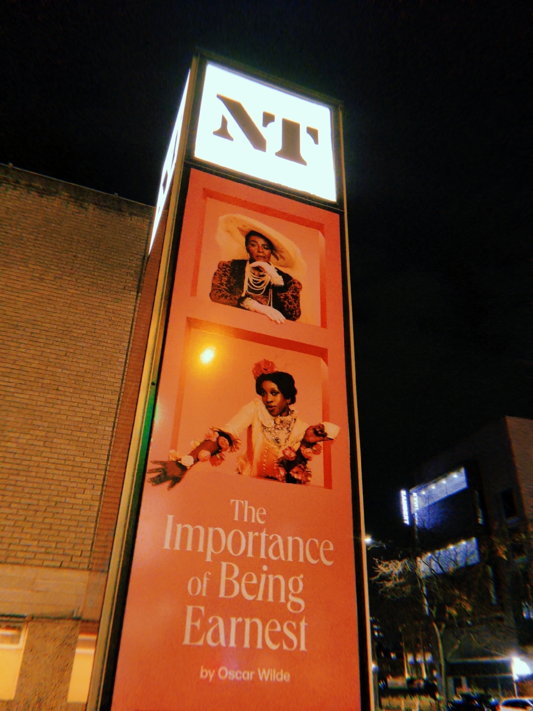 A tall outdoor advertisement board for "The Importance of Being Earnest" by Oscar Wilde is illuminated at night, featuring Sharon D Clarke and Ronkẹ Adékọluẹ́jọ́ in vintage costumes.