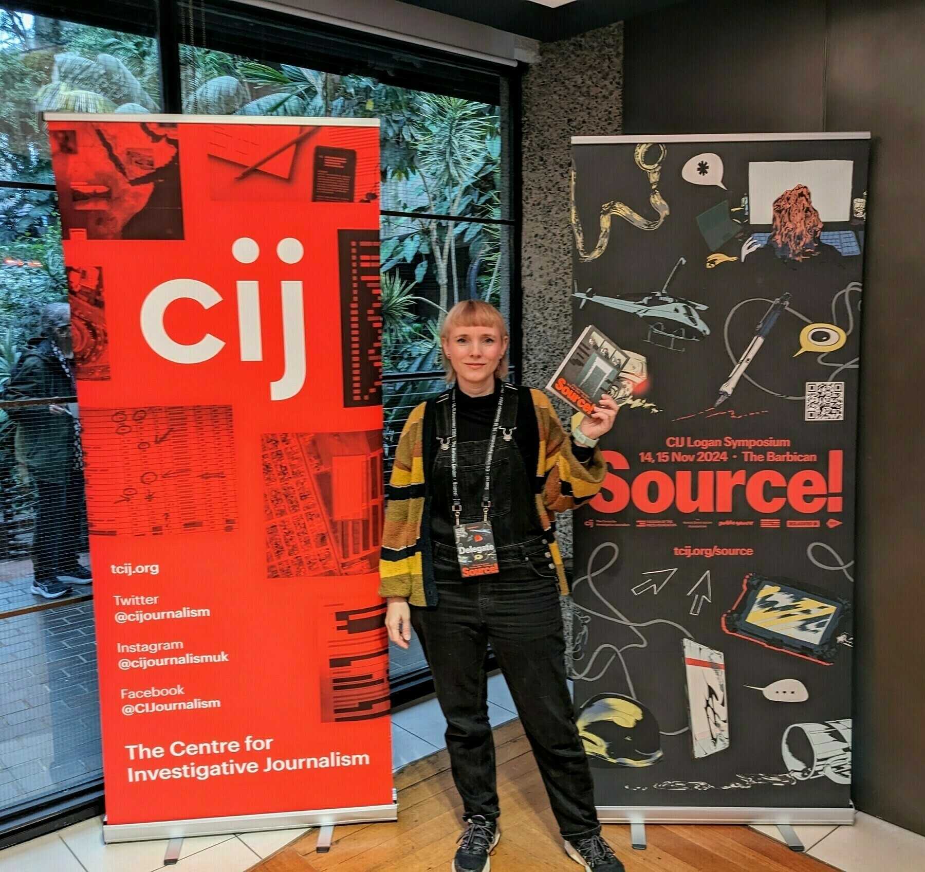 Tansy stands between two promotional banners for the Centre for Investigative Journalism, holding a programme and wearing a conference badge.