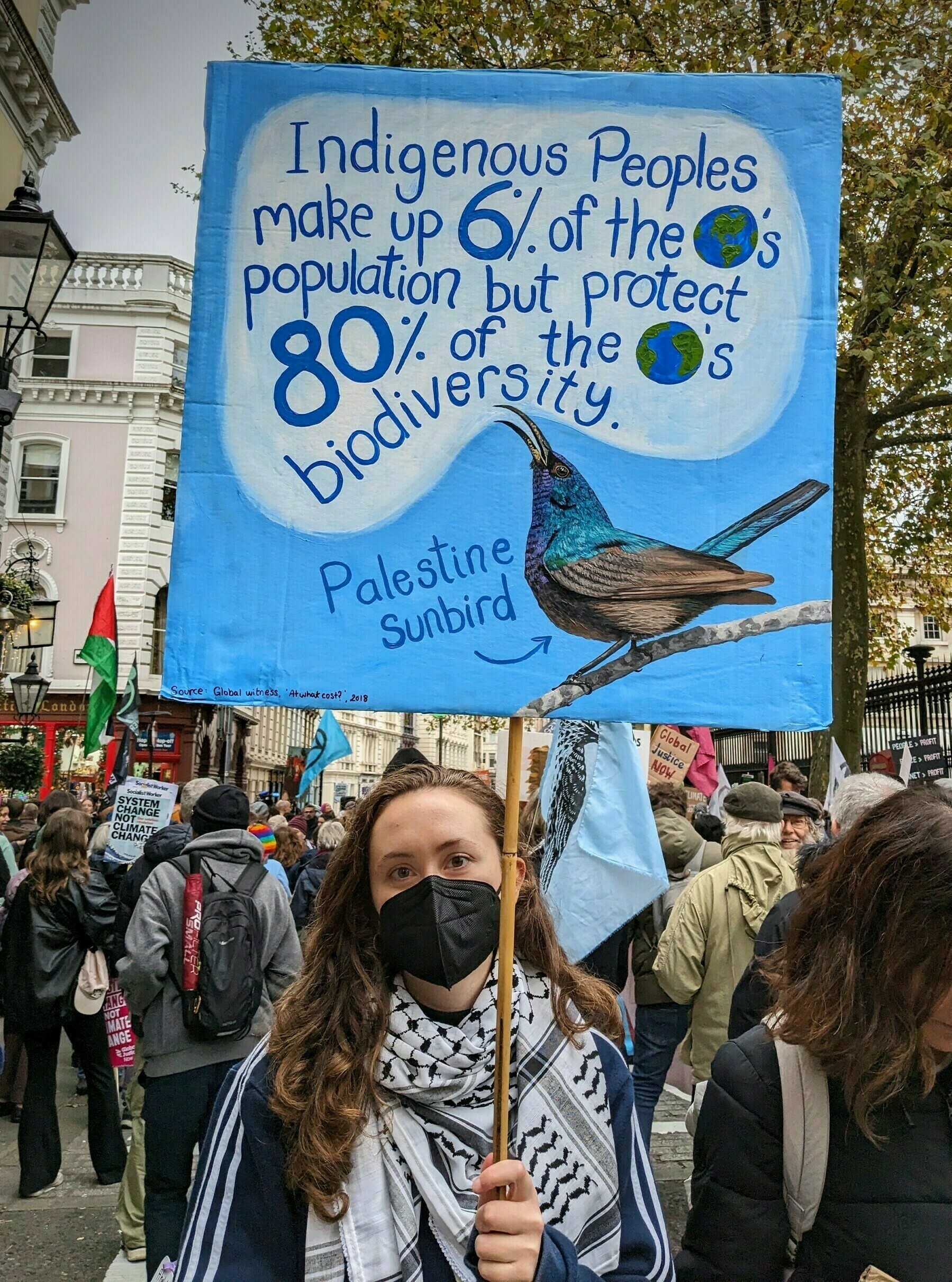 A protester holds a sign highlighting the role of Indigenous peoples in protecting biodiversity, featuring an illustration of a Palestine sunbird.