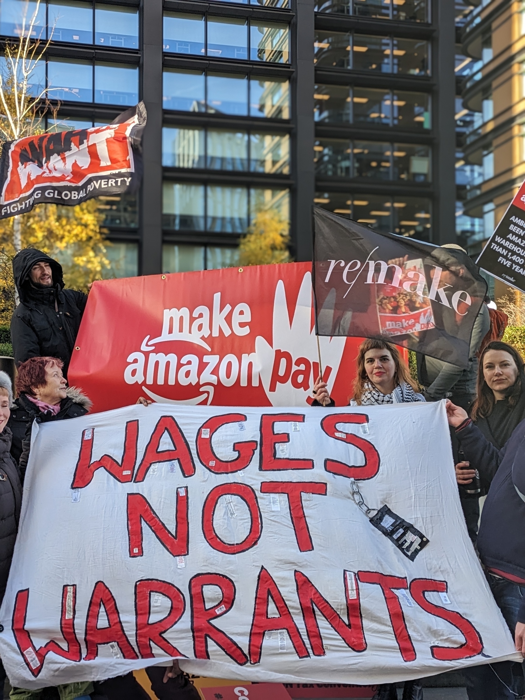 A group of people gathered to protest outside Amazon HQ. Two red and white banners say Make Amazon Pay and Wages Not Warrants