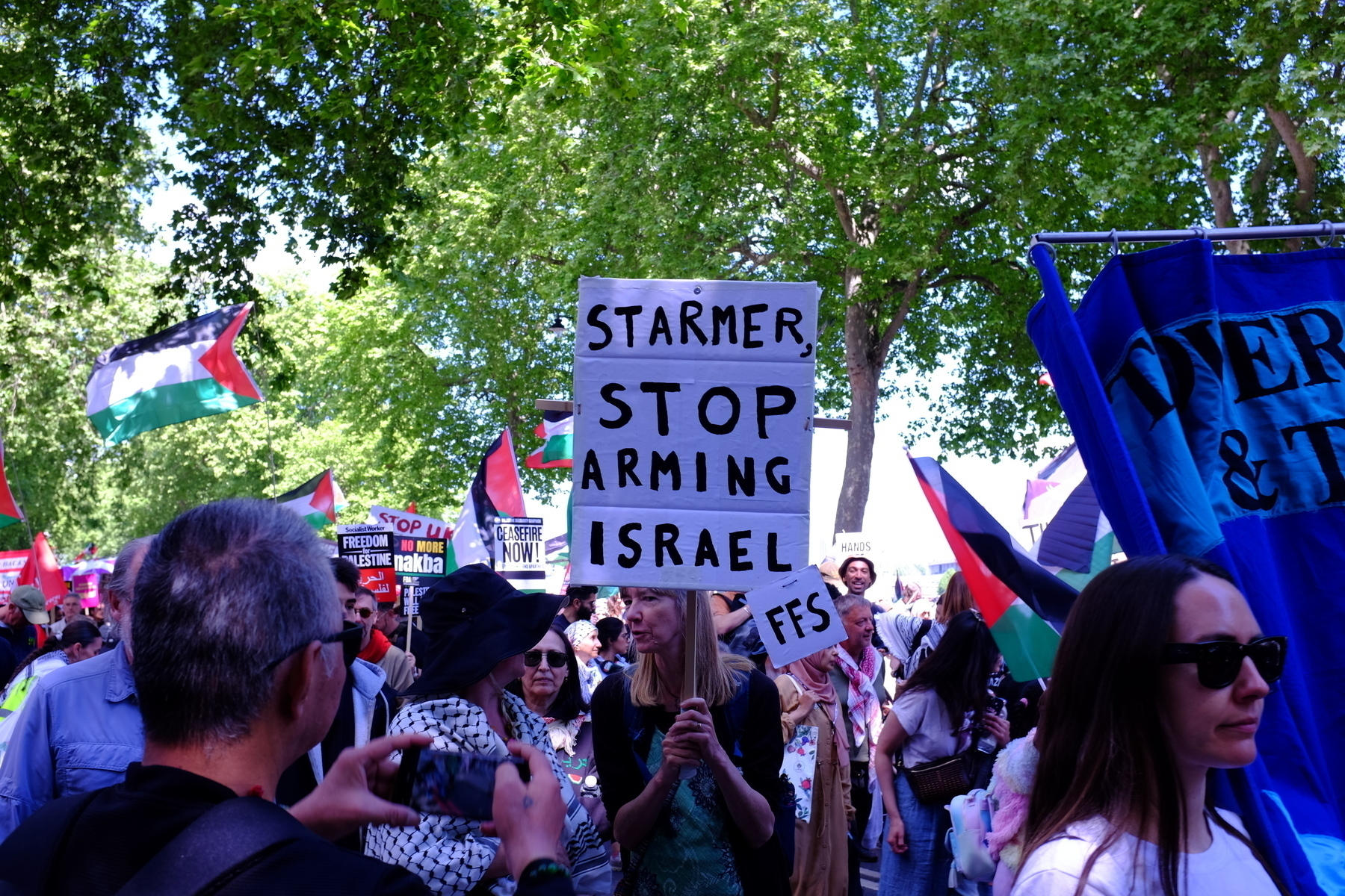 A group of people are gathered at a protest holding signs, including one that says "Starmer Stop Arming Israel FFS," surrounded by Palestinian flags.