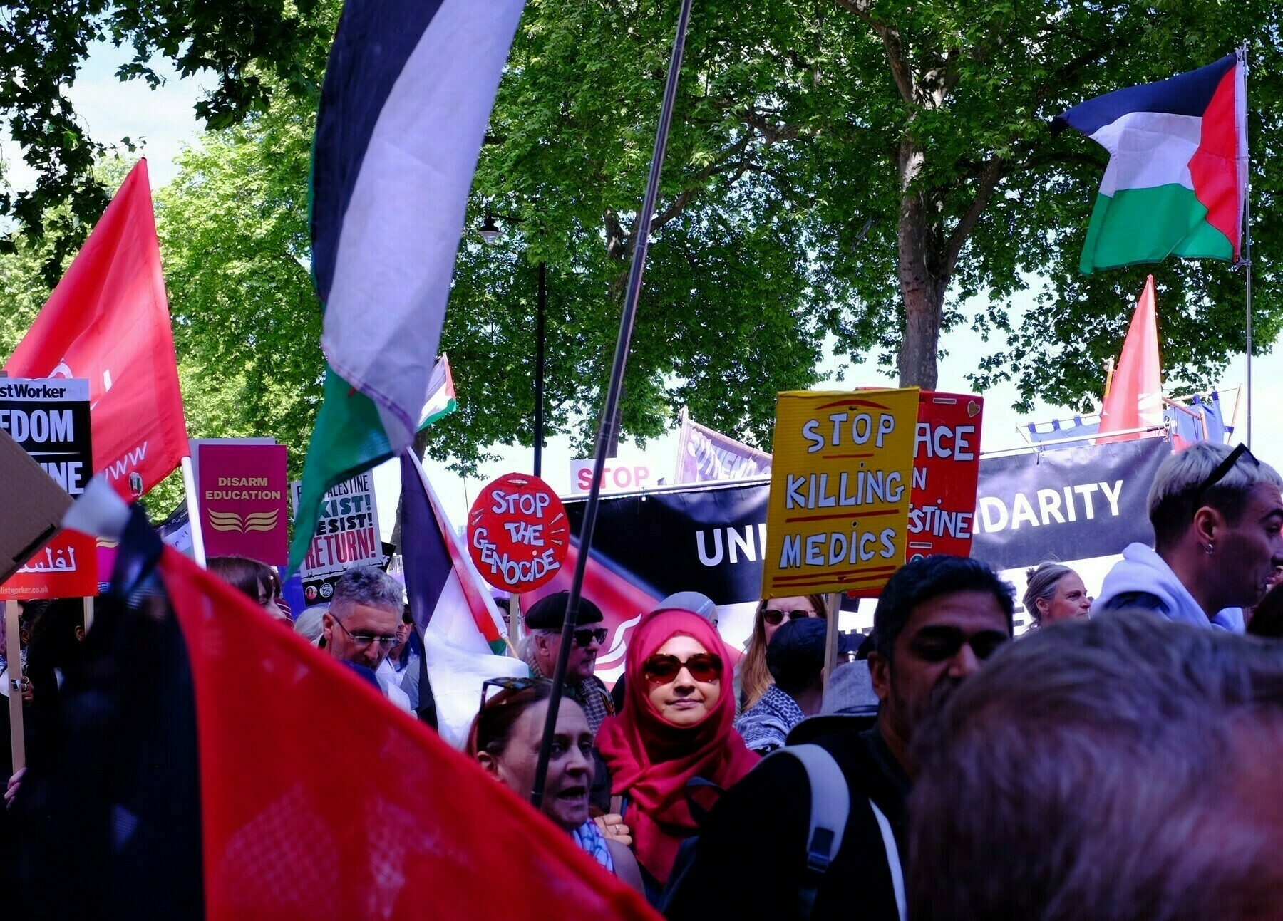 Amidst a large crowd, a woman in a red hijab and sunglasses holds a sign that reads 'Stop Killing Medics'