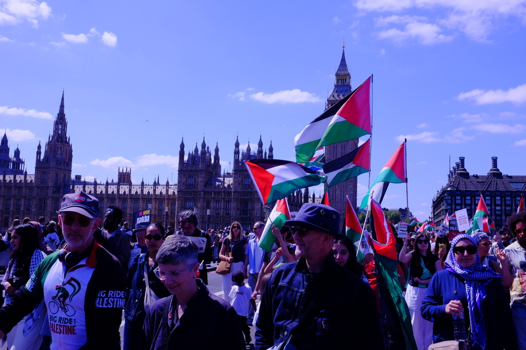 A large group of people are crossing Westminster Bridge, participating in a demonstration with Palestinian flags. In the background is London's Houses of Parliament.